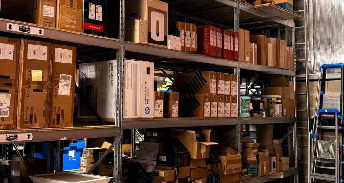 Une allée de rayonnage chargée de cartons : écrans, ordinateurs portables, imprimantes, téléphones et accessoires réseau.