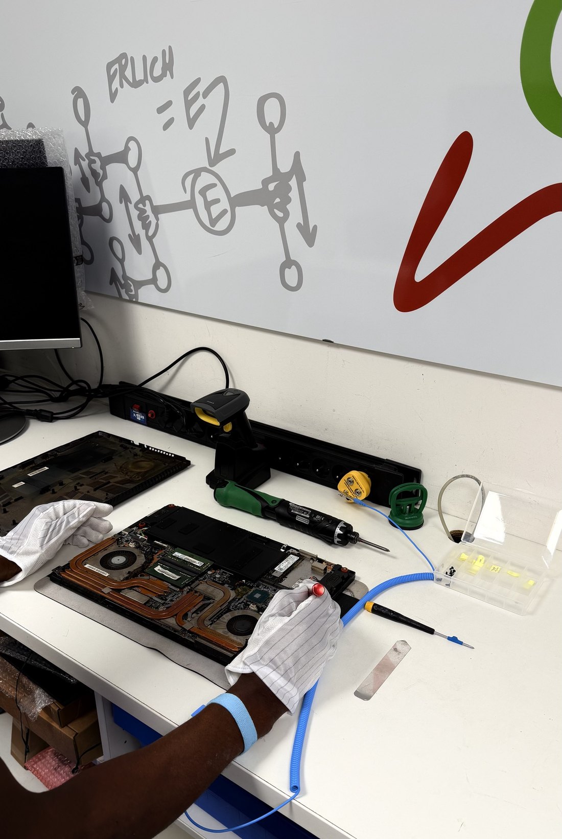Un établi d’atelier sous un panneau mural Buro24 : un ordinateur portable ouvert sur tapis antistatique, un tournevis électrique, une boîte à vis et un technicien en gants et bracelet antistatiques.