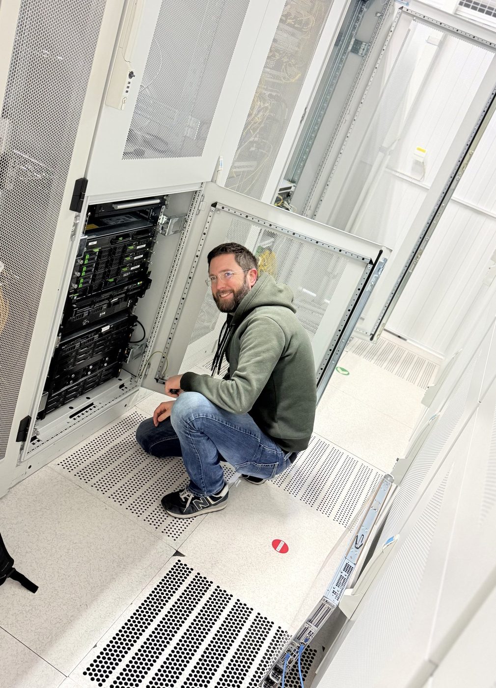 Un technicien FCC accroupi devant une baie ouverte, dans une salle informatique à faux plancher.